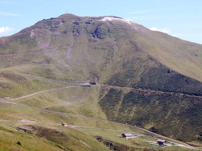 A wide landscape shot of a large, grassy mountain slope with a winding dirt road, small wooden huts at the base, and a summit featuring a cross and a patch of snow.