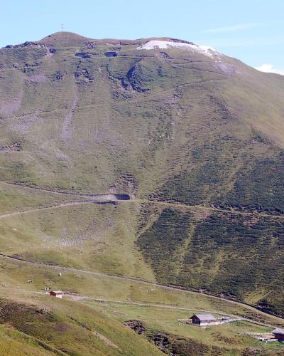 A wide landscape shot of a large, grassy mountain slope with a winding dirt road, small wooden huts at the base, and a summit featuring a cross and a patch of snow.