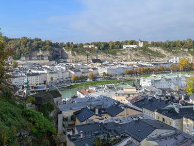 A high-angle panoramic view of the city of Salzburg, Austria, looking across the Salzach River towards the historic center, the Hohensalzburg Fortress, and the Kapuzinerberg hill in the background.