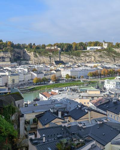 A high-angle panoramic view of the city of Salzburg, Austria, looking across the Salzach River towards the historic center, the Hohensalzburg Fortress, and the Kapuzinerberg hill in the background.