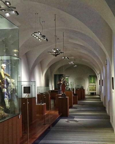 A long, vaulted gallery hallway within a museum, featuring glass display cases with ornate religious sculptures on the left, wooden pedestals, and framed artworks on the right wall, under track lighting.