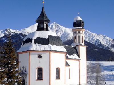 A white baroque church with a distinctive octagonal dome and a separate bell tower, covered in snow with mountains in the background.