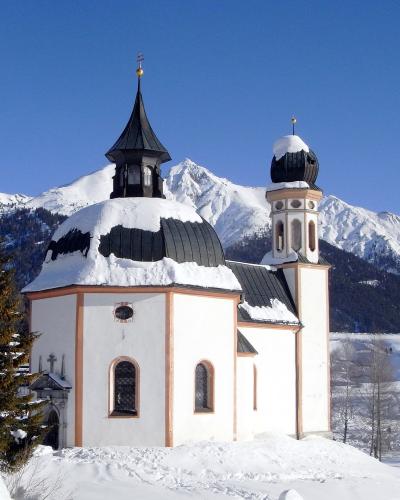 A white baroque church with a distinctive octagonal dome and a separate bell tower, covered in snow with mountains in the background.