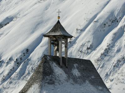 A small, white-walled chapel with a wooden shingle roof and an open wooden bell tower topped with a cross, set against a steep, snow-covered mountainside.