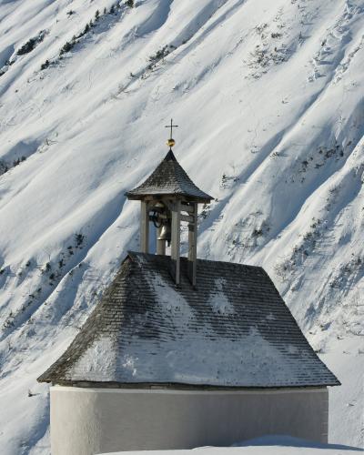 A small, white-walled chapel with a wooden shingle roof and an open wooden bell tower topped with a cross, set against a steep, snow-covered mountainside.