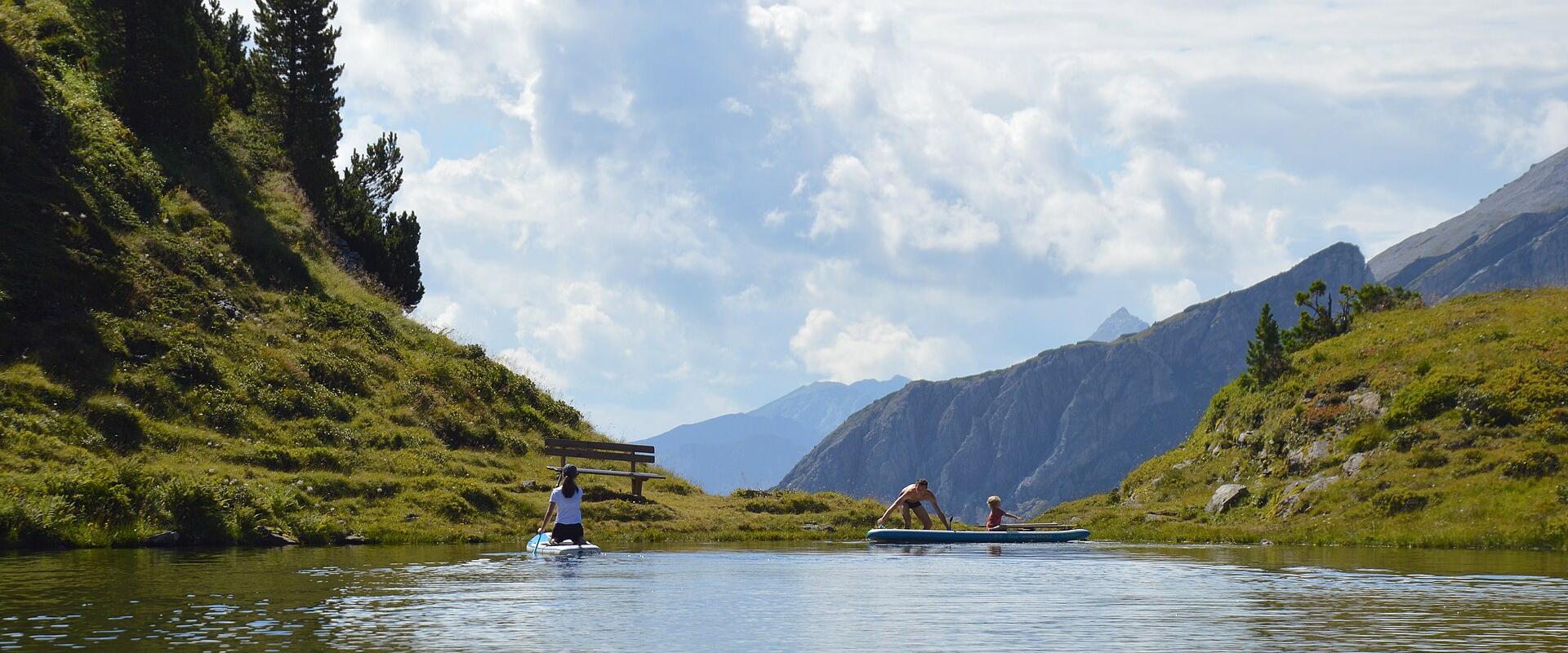 The image shows three people on paddleboards on a calm alpine lake surrounded by grassy slopes and coniferous trees, with rugged mountain peaks in the background under a partly cloudy sky; no signage or text is visible.