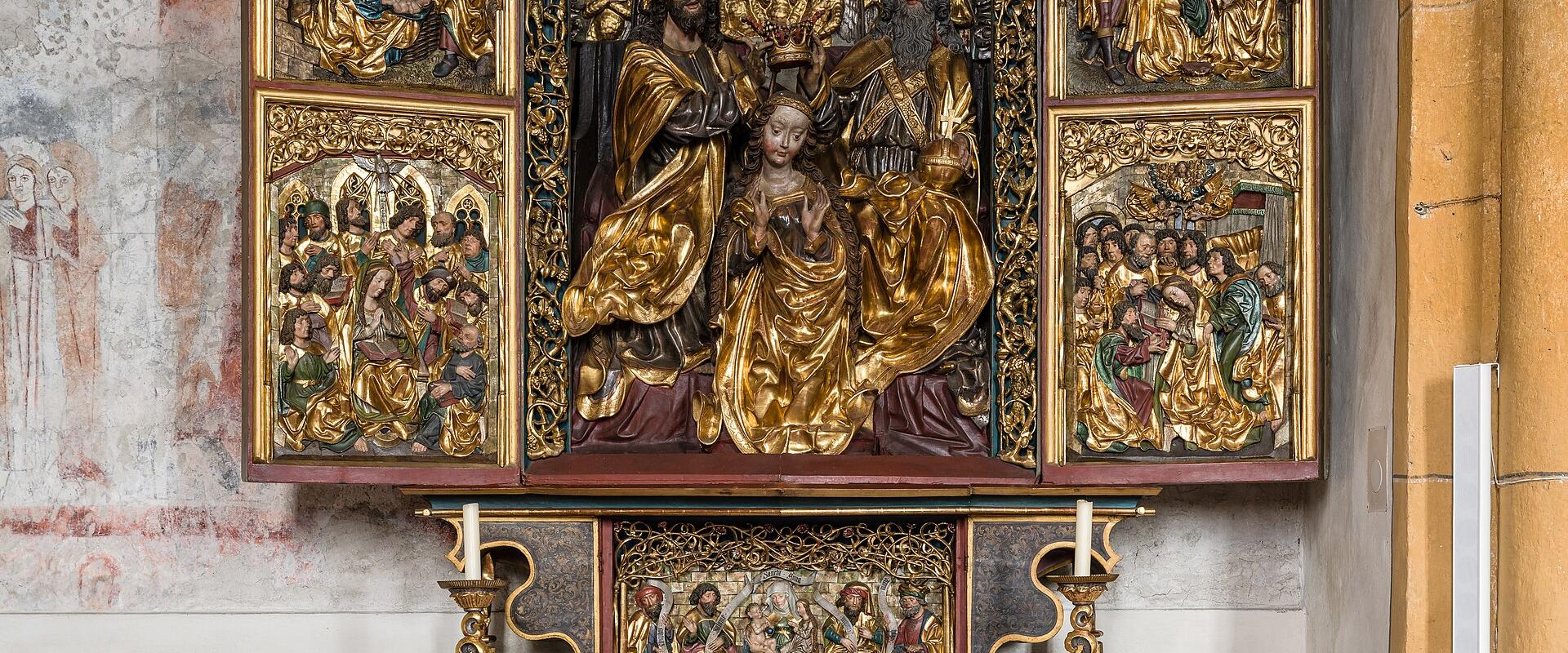 The image shows an ornate, gilded wooden altarpiece (Flügelaltar) with carved religious figures and painted panels, installed above a white altar cloth in a church interior with faded frescoes on the wall.