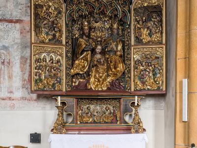 The image shows an ornate, gilded wooden altarpiece (Flügelaltar) with carved religious figures and painted panels, installed above a white altar cloth in a church interior with faded frescoes on the wall.