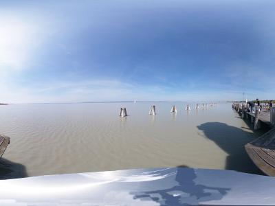 A 360-degree panoramic view of a wooden pier extending into a large, calm lake, framed by two white cylindrical lighthouse towers with red horizontal stripes on either side.