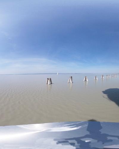 A 360-degree panoramic view of a wooden pier extending into a large, calm lake, framed by two white cylindrical lighthouse towers with red horizontal stripes on either side.