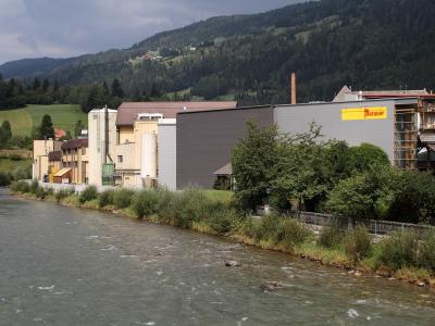 An industrial brewery complex situated on the bank of a river with forested hills in the background, featuring a prominent yellow sign on the building.