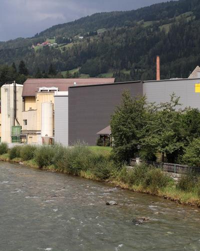 An industrial brewery complex situated on the bank of a river with forested hills in the background, featuring a prominent yellow sign on the building.