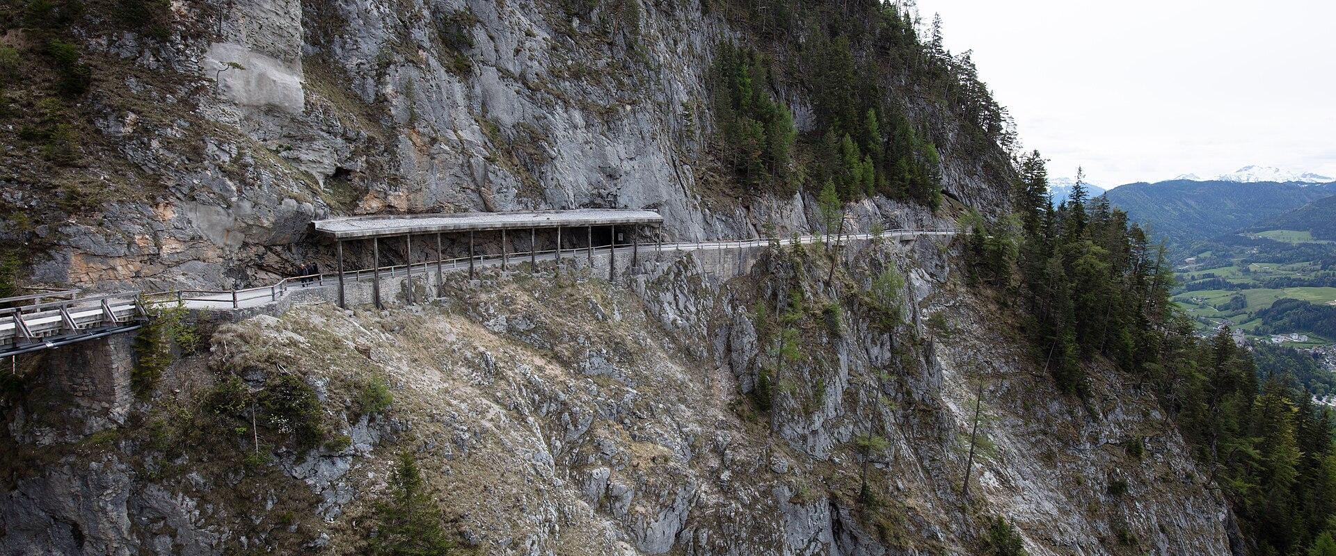 A wooden walkway with a roofed shelter section is built along a steep, rocky mountainside, with forested slopes and distant green valleys visible in the background.