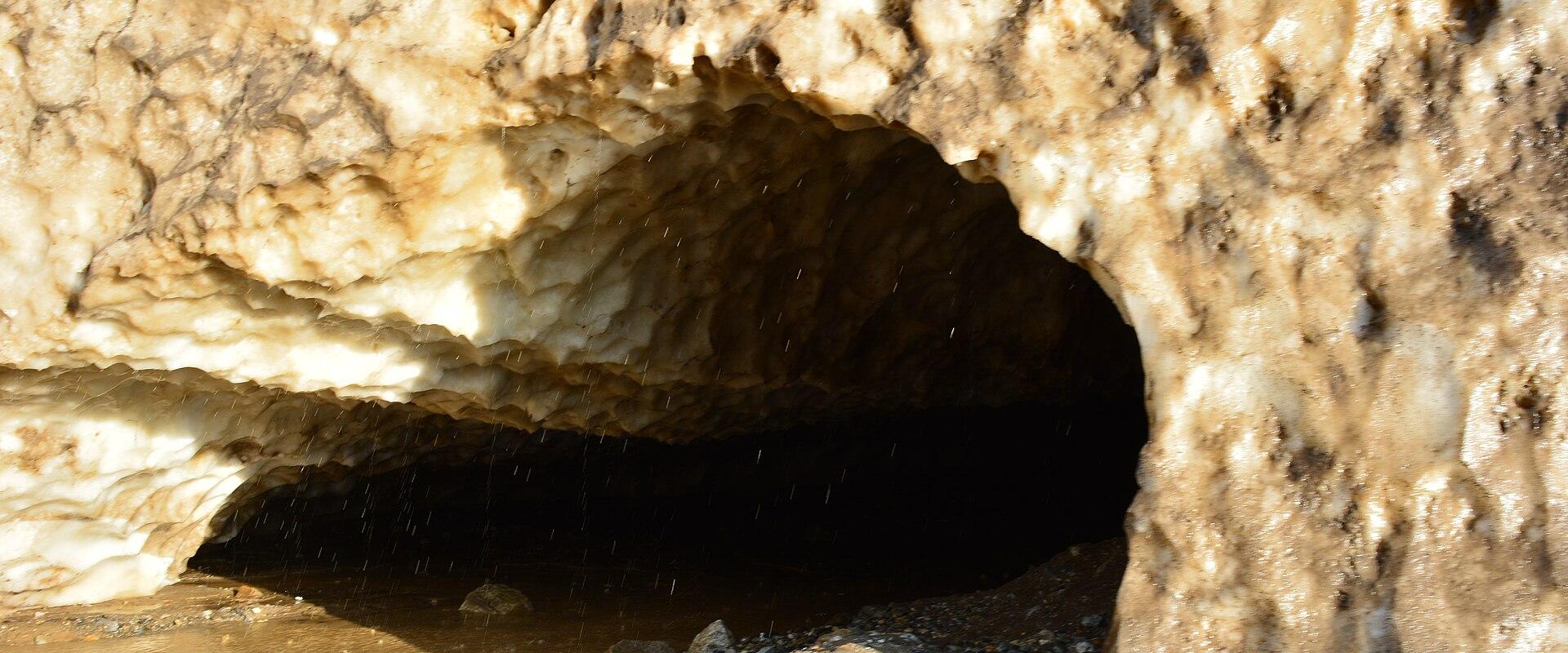 A close-up view of a melting ice cave entrance with dirty, textured ice walls, a dark interior, and meltwater pooling at the base.