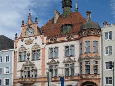 A multi-story historic building with a pink and white facade, a clock tower, a green spire, and flags flying in front, situated in a town square.