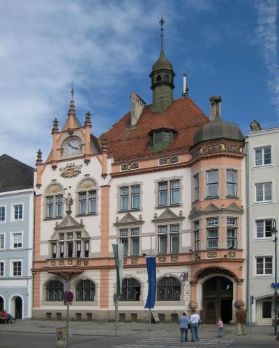 A multi-story historic building with a pink and white facade, a clock tower, a green spire, and flags flying in front, situated in a town square.
