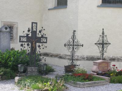 A section of a cemetery with three ornate metal crosses and grave markers set against the white stucco wall of a building.