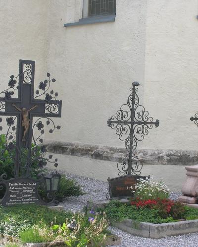 A section of a cemetery with three ornate metal crosses and grave markers set against the white stucco wall of a building.