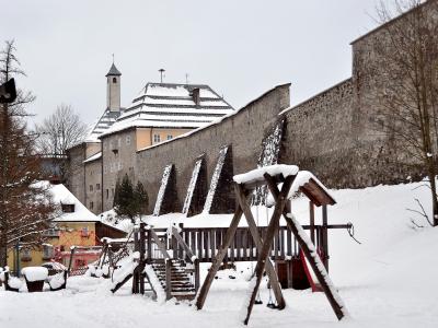 A snow-covered wooden playground structure in the foreground with a tall stone fortification wall and a building with a tower in the background.