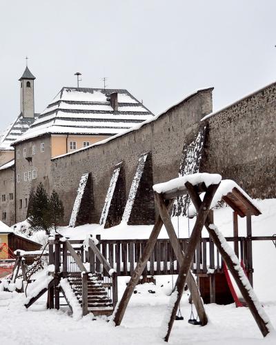 A snow-covered wooden playground structure in the foreground with a tall stone fortification wall and a building with a tower in the background.