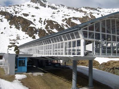 A long, elevated glass-enclosed walkway or station structure supported by pillars, with a blue booth and cable car cabins visible underneath, set against a snowy mountain backdrop.