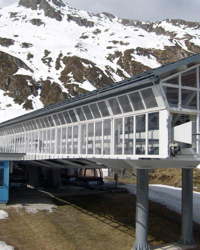 A long, elevated glass-enclosed walkway or station structure supported by pillars, with a blue booth and cable car cabins visible underneath, set against a snowy mountain backdrop.