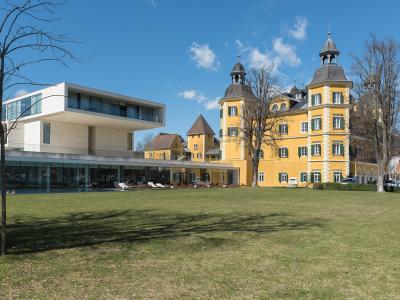 A yellow Baroque-style palace with distinctive corner towers and green shutters stands next to a modern white glass building, set behind a large grassy lawn under a blue sky.