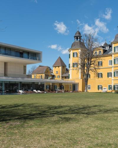 A yellow Baroque-style palace with distinctive corner towers and green shutters stands next to a modern white glass building, set behind a large grassy lawn under a blue sky.