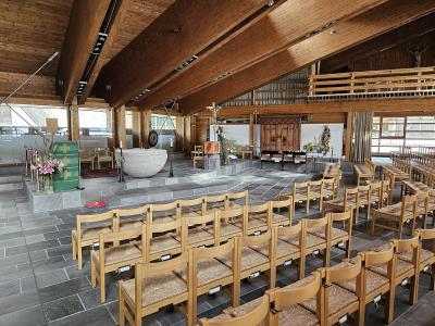 The interior of a modern church featuring wooden ceiling beams, stone flooring, rows of wooden chairs, and a large stone baptismal font.