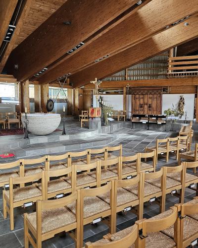 The interior of a modern church featuring wooden ceiling beams, stone flooring, rows of wooden chairs, and a large stone baptismal font.