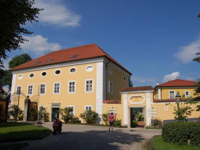 A yellow two-story building with a red tiled roof and white architectural trim, featuring an arched gateway entrance and a person walking on the gravel driveway.