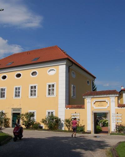 A yellow two-story building with a red tiled roof and white architectural trim, featuring an arched gateway entrance and a person walking on the gravel driveway.
