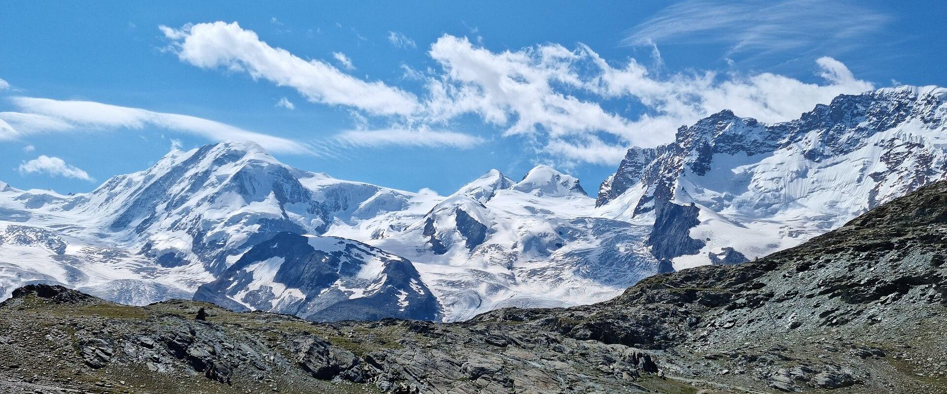 A high-altitude alpine lake (Riffelsee) with green grassy slopes in the foreground, rocky terrain, and a backdrop of snow-covered peaks under a blue sky with scattered clouds; no visible signage or text.