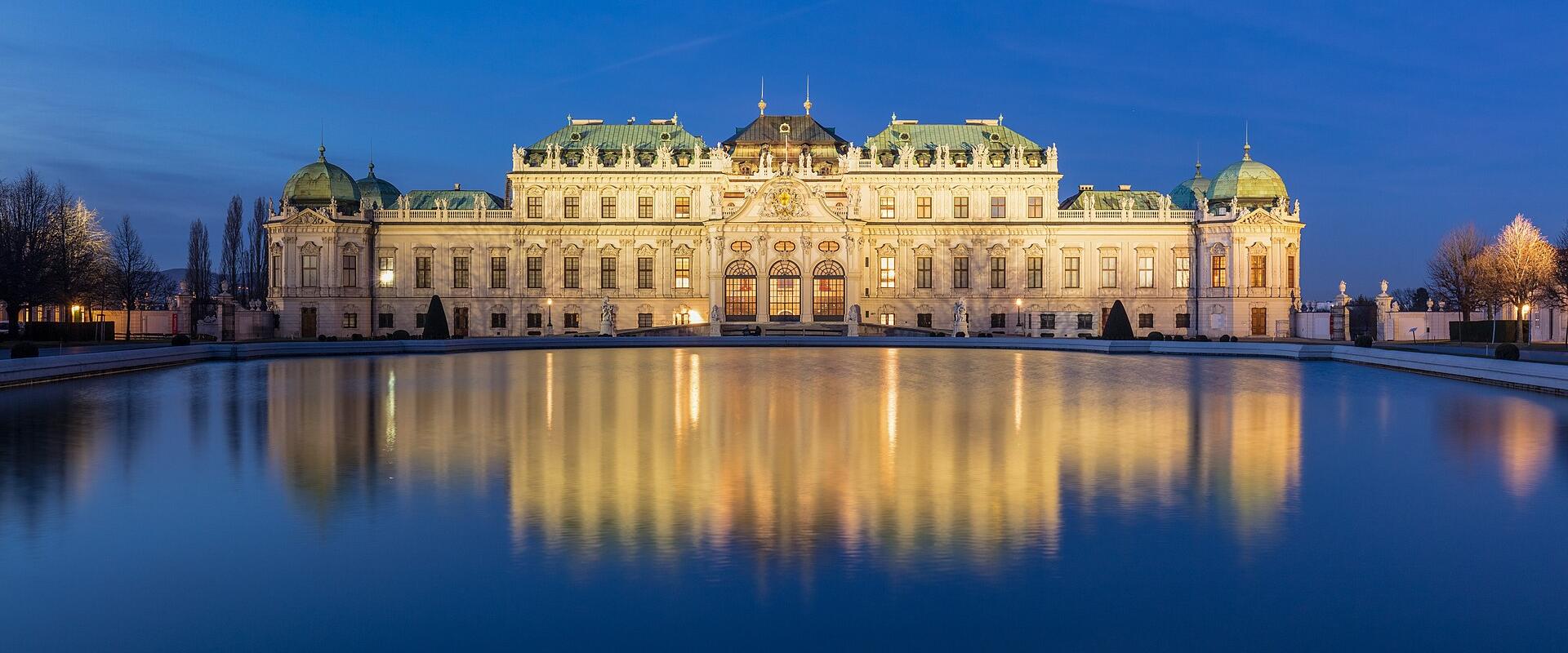 The image shows the Belvedere Palace in Vienna, Austria, illuminated at dusk with its reflection visible in a large rectangular pool of water in the foreground; the architecture features Baroque design with green copper domes and ornate facades.