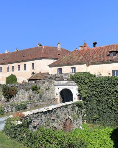 A large, light-colored castle complex with a red-tiled roof, stone fortifications, and an arched gateway, viewed from a gravel path with green vegetation and a clear blue sky.