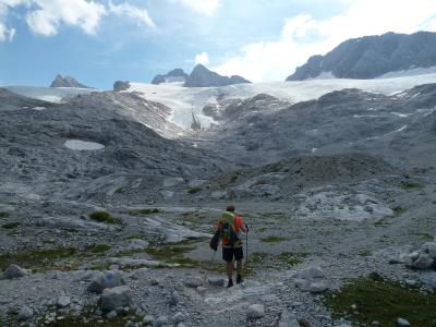 A hiker with a backpack and trekking poles walks across a rocky, moraine-covered landscape towards a large glacier tongue in the background under a partly cloudy sky.