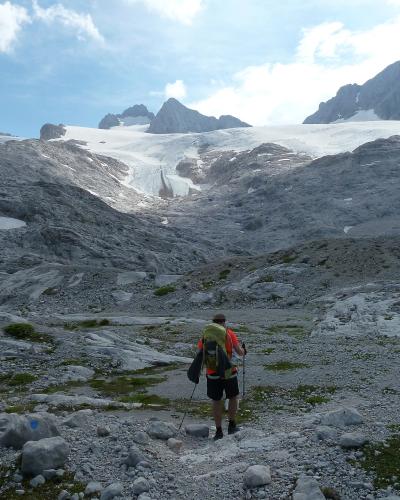 A hiker with a backpack and trekking poles walks across a rocky, moraine-covered landscape towards a large glacier tongue in the background under a partly cloudy sky.
