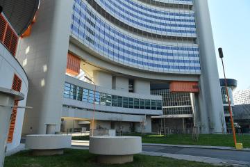 A low-angle exterior shot of a large, modernist office complex with curved white concrete facades, blue-tinted glass windows, and distinctive orange ventilation louvers, set against a clear sky with grass and concrete planters in the foreground.
