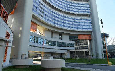 A low-angle exterior shot of a large, modernist office complex with curved white concrete facades, blue-tinted glass windows, and distinctive orange ventilation louvers, set against a clear sky with grass and concrete planters in the foreground.