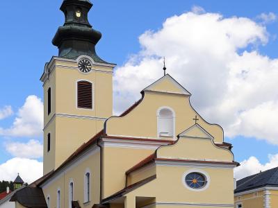 A yellow Catholic church with a tall bell tower topped by a golden cross and a dark green spire, set against a blue sky with white clouds.