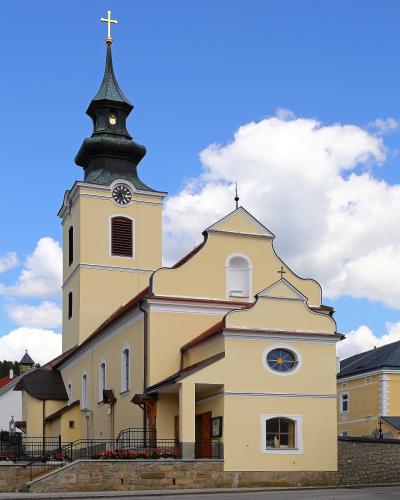 A yellow Catholic church with a tall bell tower topped by a golden cross and a dark green spire, set against a blue sky with white clouds.