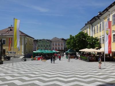 A wide, paved town square featuring a distinctive grey and white chevron-patterned pavement, flanked by yellow and green historic buildings, market stalls with green canopies, and flags including an Austrian flag.