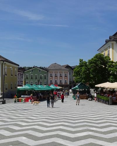 A wide, paved town square featuring a distinctive grey and white chevron-patterned pavement, flanked by yellow and green historic buildings, market stalls with green canopies, and flags including an Austrian flag.