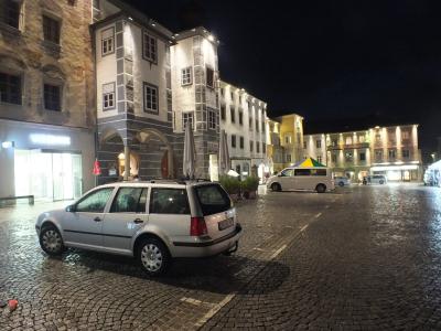 A night view of a cobblestone town square featuring a white station wagon in the foreground, historic buildings with arcades, and a white van parked further back.