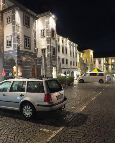 A night view of a cobblestone town square featuring a white station wagon in the foreground, historic buildings with arcades, and a white van parked further back.