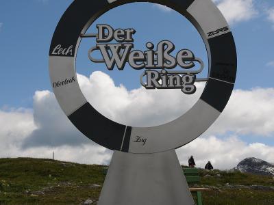 A large metal sculpture of a ring with alternating black and white segments, featuring the text "Der Weiße Ring" in the center and names of locations like "Lech," "Zürs," "Oberlech," and "Zug" on the ring, set against a grassy hillside with mountains