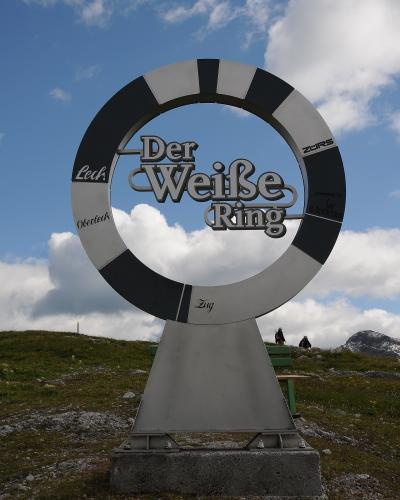 A large metal sculpture of a ring with alternating black and white segments, featuring the text "Der Weiße Ring" in the center and names of locations like "Lech," "Zürs," "Oberlech," and "Zug" on the ring, set against a grassy hillside with mountains