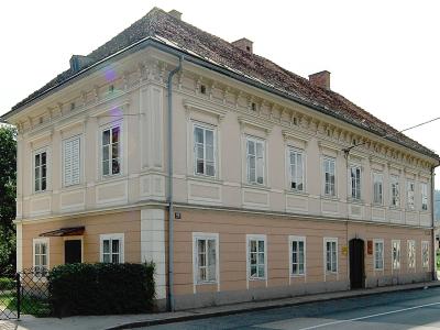 A two-story, cream-colored building with a tiled roof and white window frames, featuring a street number plaque reading "69" on the corner.