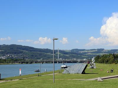 A grassy riverside park with modern sculptures, a lamp post, and a large truss bridge spanning a wide river with hills in the background.