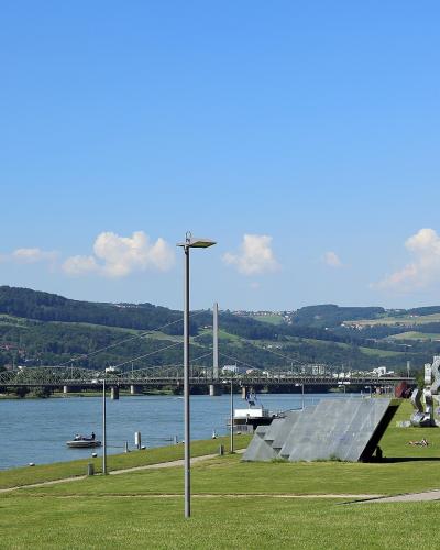A grassy riverside park with modern sculptures, a lamp post, and a large truss bridge spanning a wide river with hills in the background.